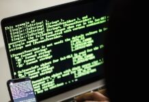 unrecognizable hacker with smartphone typing on laptop at desk